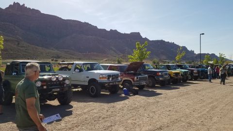 1985 Ford Bronco - A gathering of Ford Broncos and trucks parked in a lot with a mountainous backdrop.