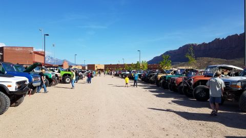 1985 Ford Bronco - A gathering of off-road vehicles are parked in a lot under a clear blue sky with mountains in the distance.
