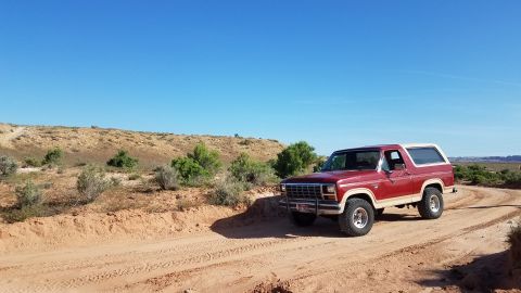 1985 Ford Bronco - This red 1985 Ford Bronco with a white top is seen driving on a dusty trail in a desert landscape.