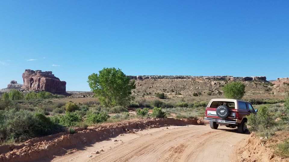Red and tan Ford Bronco with a spare tire on the back, driving on a dirt road in a desert landscape