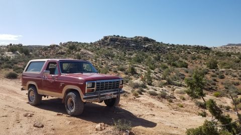 1985 Ford Bronco - A red Ford Bronco is parked on a dirt road in a desert landscape with rocky hills.