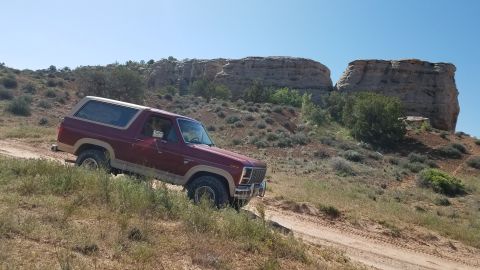 1985 Ford Bronco - 1985 Ford Bronco navigating a dusty trail through a desert landscape under a clear blue sky.