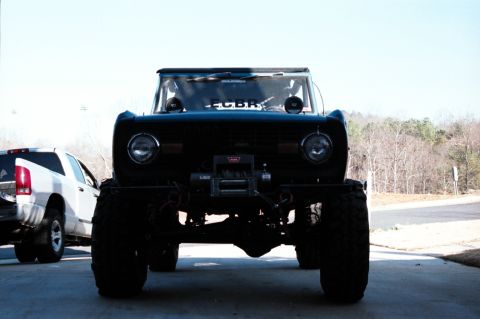 1976 Ford Bronco - A heavily modified black SUV with a winch is parked in front of a white SUV.