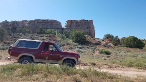 1985 Ford Bronco - A red Ford Bronco sits on a dirt road with large rock formations and sparse vegetation under a clear blue sky.