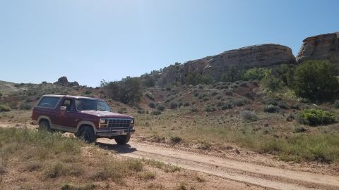 1985 Ford Bronco - A 1985 Ford Bronco navigates a dirt track through a dry, hilly landscape under a clear blue sky.