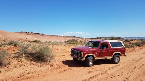 1985 Ford Bronco - A classic red Ford Bronco is parked on a sandy trail in a desert environment with red rock formations in the background.