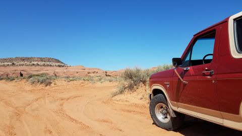 1985 Ford Bronco - A red and tan Ford Bronco is parked on a dirt road in a desert landscape with red rock formations.