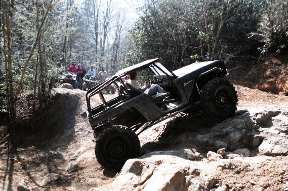 Black 1976 Ford Bronco climbing rocks on a wooded trail, with people on ATVs in the background