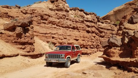 1985 Ford Bronco - A red 1985 Ford Bronco navigates a dusty trail between towering red rock formations under a clear blue sky.