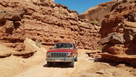1985 Ford Bronco - 1985 Ford Bronco navigates a narrow dirt path between towering red rock formations under a clear blue sky.