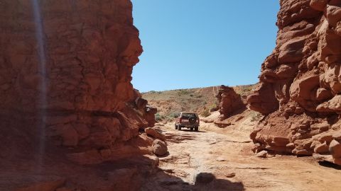 1985 Ford Bronco - A red SUV navigates a narrow dirt path flanked by towering red rock cliffs under a clear blue sky.