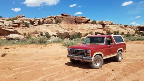 1985 Ford Bronco - A red Ford Bronco is parked on a dusty trail in a desert landscape with sandstone formations under a blue sky.