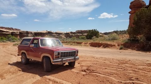 1985 Ford Bronco - A red Ford Bronco is parked on a dusty trail in a desert landscape under a blue sky with scattered clouds.