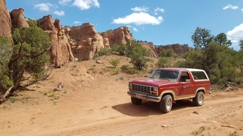 1985 Ford Bronco - Red Ford Bronco on a dirt road with red rock formations in the background.