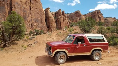 1985 Ford Bronco - A red Ford Bronco with a tan top is parked in a desert landscape with large rock formations.