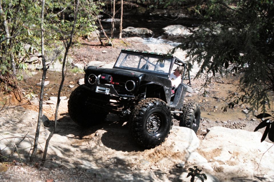 Black 1976 Ford Bronco modified for off-roading, driving over rocks near a stream