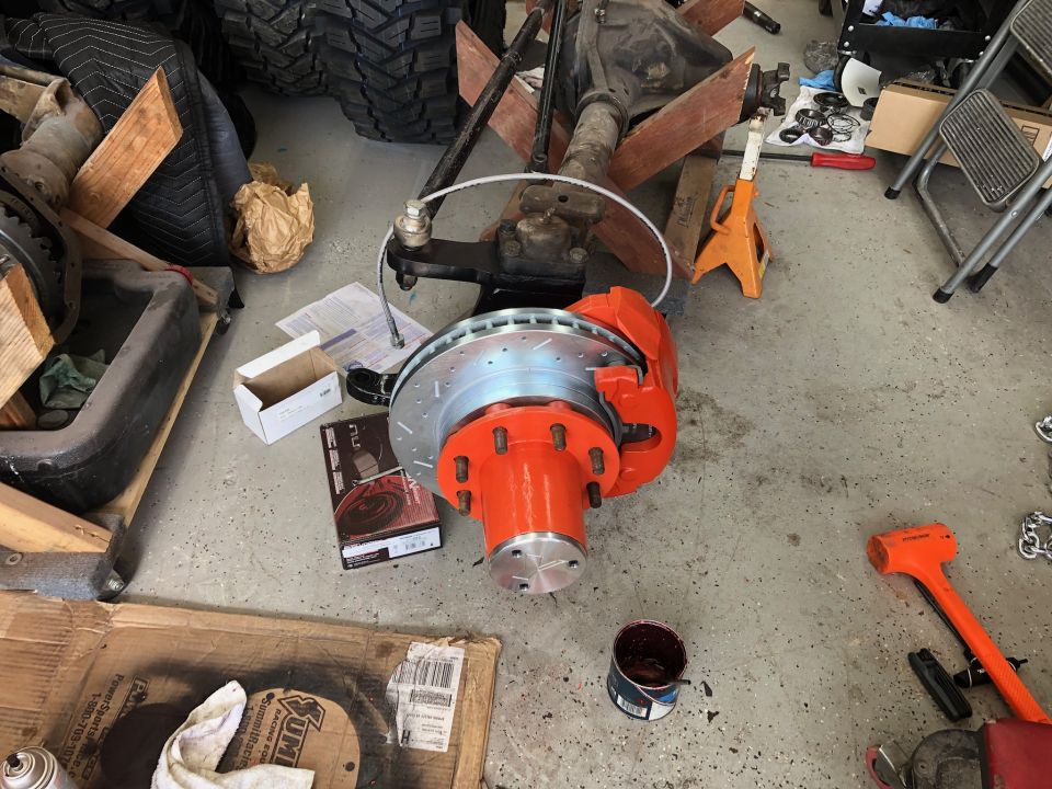 Front axle assembly with orange brake calipers and drilled rotors. Tires and other parts are in the background