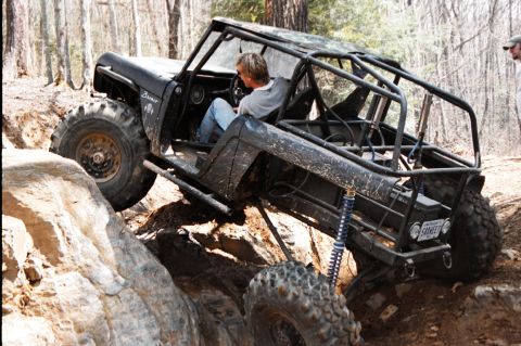 1976 Ford Bronco - Chopped black Ford Bronco rock buggy flexing over a rock ledge on a wooded trail, seen from the driver's side rear with the driver seated a…