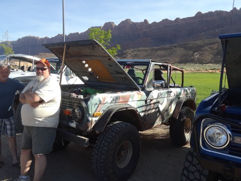 1985 Ford Bronco - A camouflaged Ford Bronco is parked next to another vehicle, with its hood open under a clear sky.