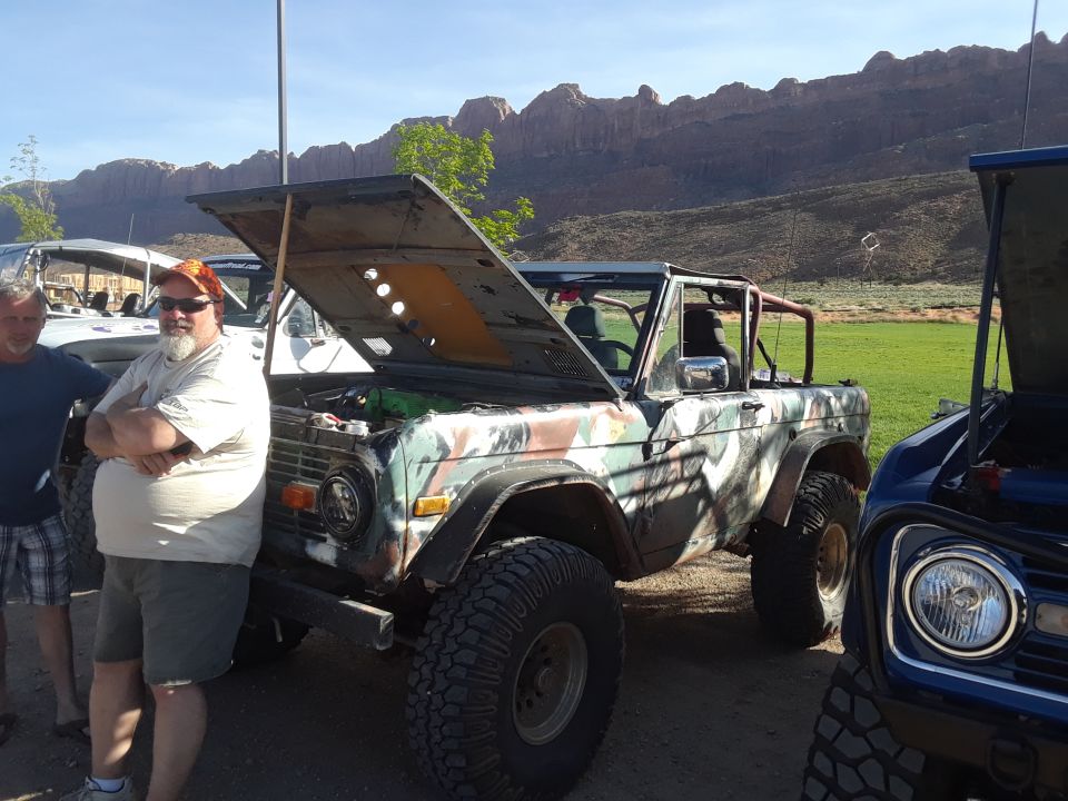 Camouflaged Ford Bronco with hood open, large tires, and a man standing beside it