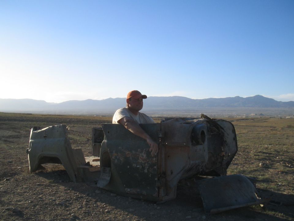 man in orange cap sits in a rusted vehicle body, desert landscape behind