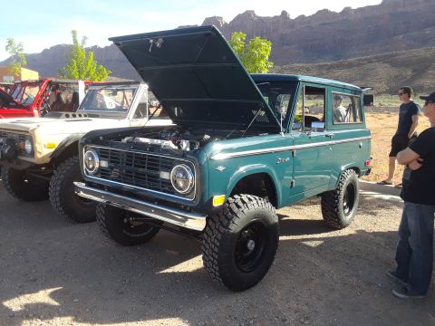 1985 Ford Bronco - A green 1985 Ford Bronco, hood raised to reveal the engine, sits parked alongside other vehicles in a desert landscape.