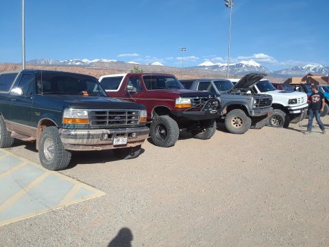 1985 Ford Bronco - A lineup of Ford Bronco SUVs are parked outdoors under a clear blue sky with mountains in the distance.