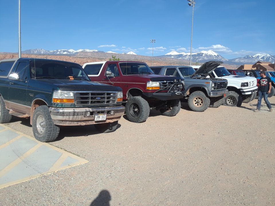 Several Ford Bronco SUVs parked in a lot with mountains in the background. One has its hood up