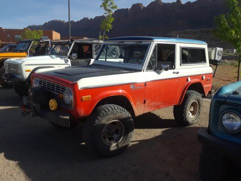 1985 Ford Bronco - A red and white Ford Bronco is parked in a lot with other vehicles and mountains in the background.