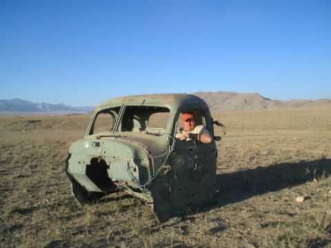 1981 Ford Bronco - A person sits in the driver's seat of a heavily damaged, old truck cab abandoned in a desolate landscape.