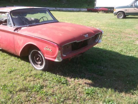 1960 Ford Starliner - This red 1960 Ford Starliner is missing its grille and shows signs of wear, parked in a grassy area with other vehicles visible.