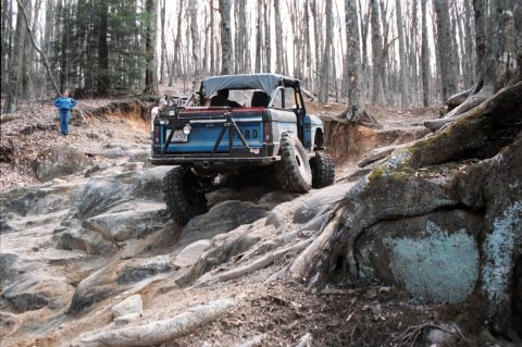 1976 Ford Bronco - A blue 1976 Ford Bronco navigates a challenging rocky terrain in a wooded area.