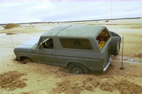 1979 Ford Bronco - A gray 1978 Ford Bronco is partially buried in sand, with a person attempting to free it.
