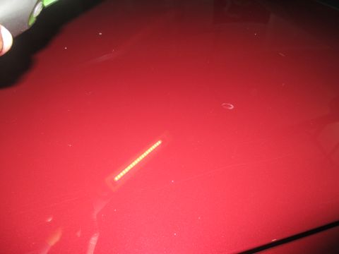 2004 Ford Crown Victoria - Close-up of a red car's hood shows surface imperfections and a light source reflection.