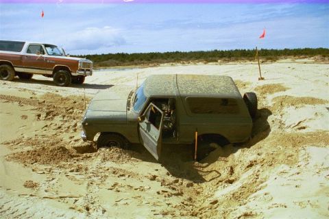 1979 Ford Bronco - Two SUVs are on a sandy terrain, one deeply stuck in a hole with its door open.