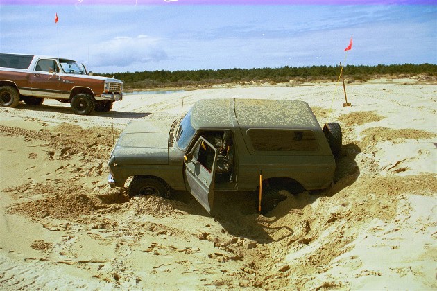 Olive green SUV with open driver door stuck in sand, another brown SUV in background