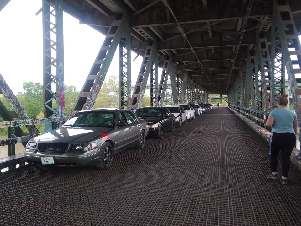Gray Ford Crown Victoria in a line of cars on a bridge. Red stripe on hood