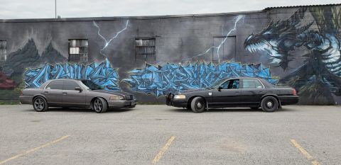 2004 Ford Crown Victoria - Two Ford Crown Victorias are parked in front of a building with a large, colorful mural.