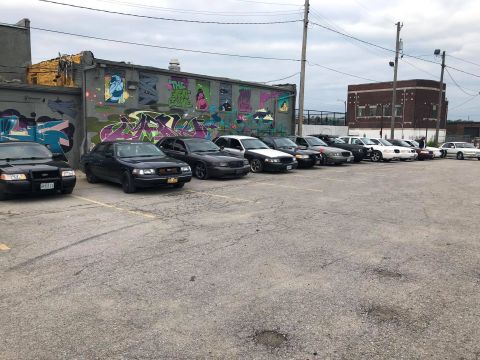 2004 Ford Crown Victoria - A row of police interceptor sedans are parked in front of a graffiti-covered building under a cloudy sky.