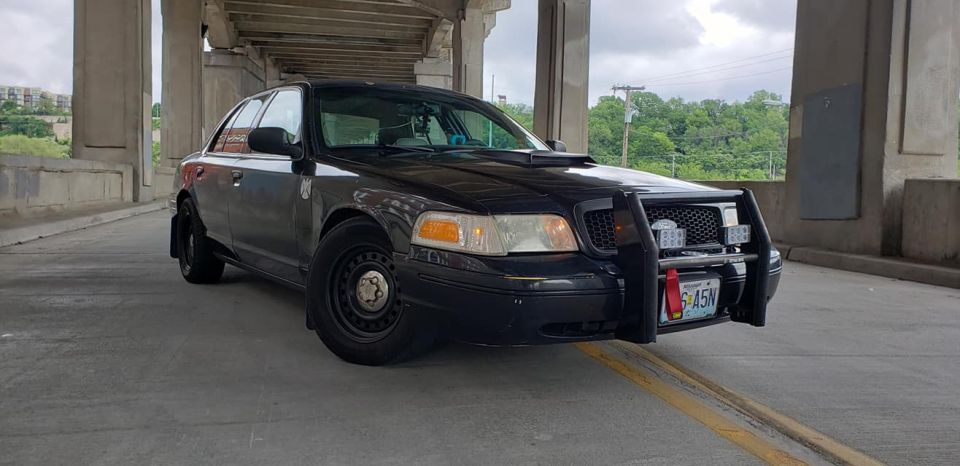 Black 2004 Ford Crown Victoria with a push bar and auxiliary lights, parked under a concrete overpass