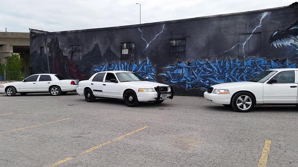 Three white Ford Crown Victorias parked in front of a mural