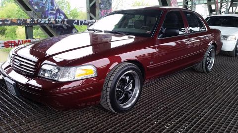 2004 Ford Crown Victoria - A dark red sedan with aftermarket wheels is parked on a metal grate under a bridge, with another white sedan visible behind it.