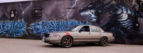 2004 Ford Crown Victoria - A tan 2004 Ford Crown Victoria with red racing stripes is parked in front of a mural a dragon and graffiti.