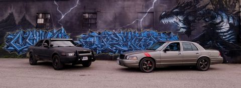 2004 Ford Crown Victoria - Two Ford Crown Victorias, one modified for off-road and the other a street build, are parked side-by-side in front of a mural.
