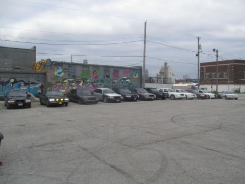 2004 Ford Crown Victoria - A row of sedans are parked in an asphalt lot with a graffiti-covered building in the background.