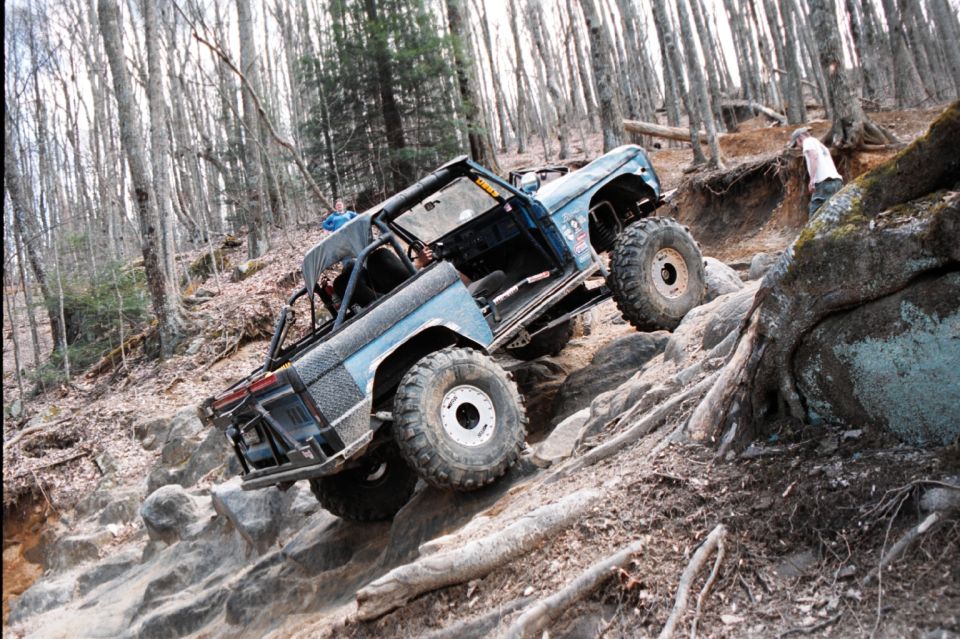 1976 Ford Bronco climbing a rocky hill in a forest, with a person standing on the side