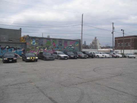 2004 Ford Crown Victoria - A fleet of sedans, predominantly Ford Crown Victorias, are parked in an urban lot under a cloudy sky.