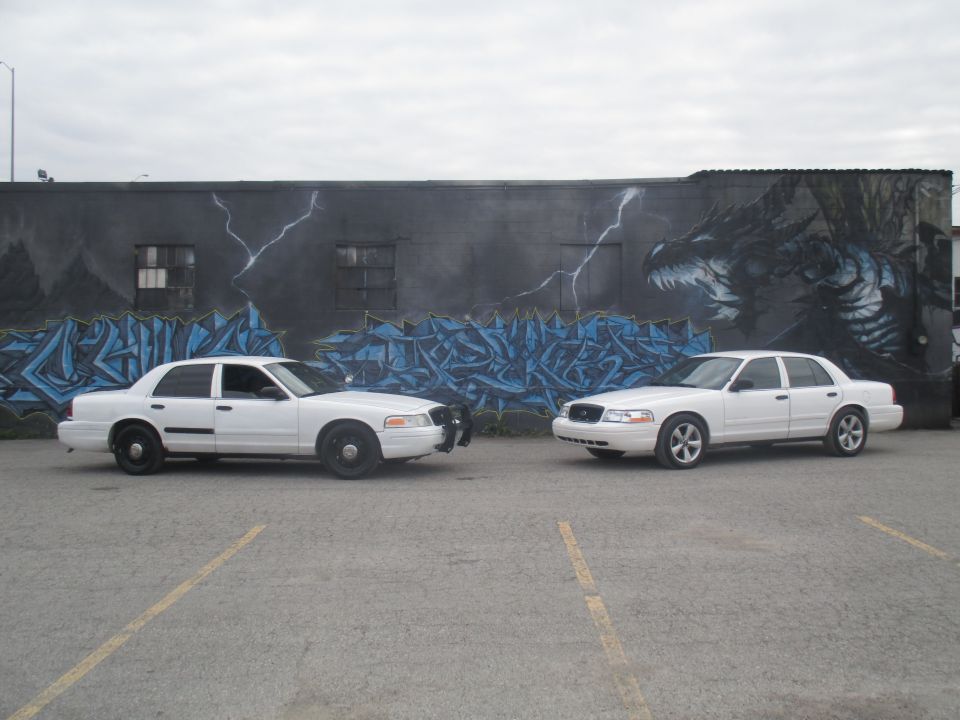 Two white 2004 Ford Crown Victoria police cars parked side-by-side in front of a graffiti mural depicting a dragon