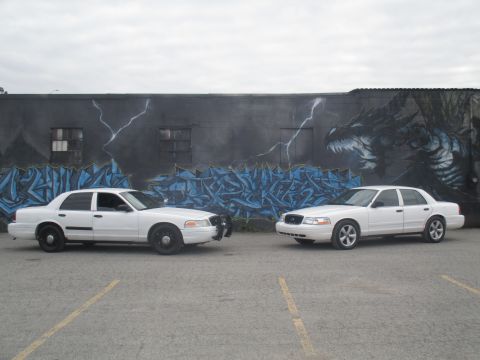2004 Ford Crown Victoria - Two white police sedans are parked side-by-side in front of a wall with graffiti art.