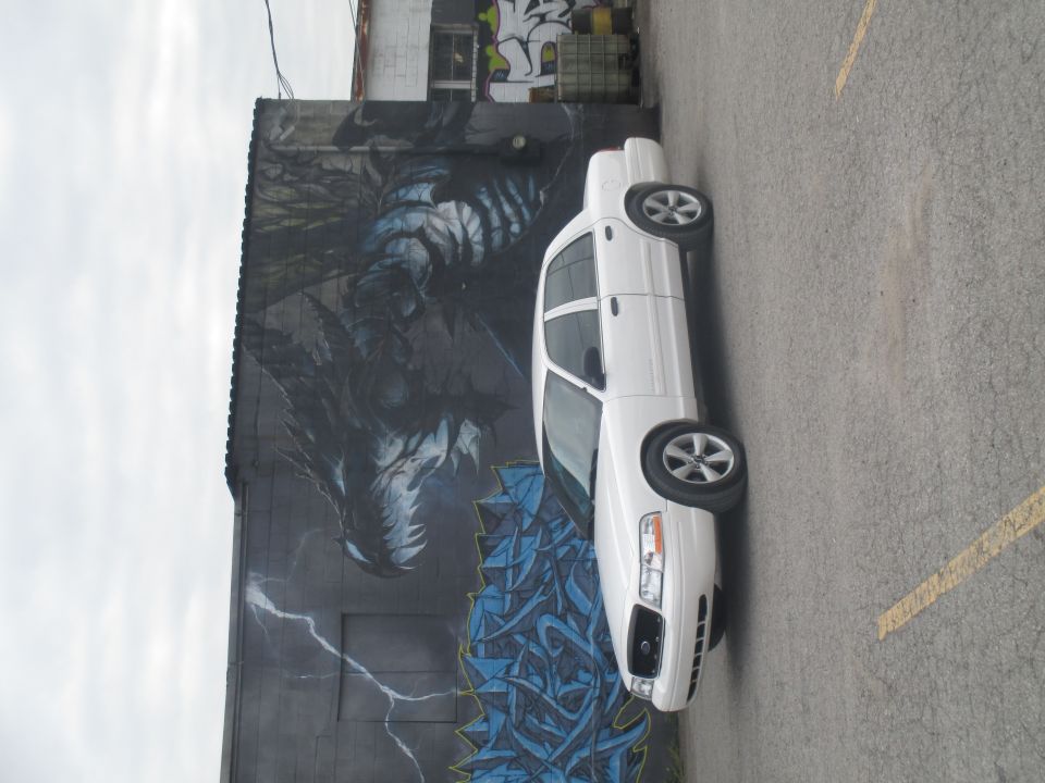 White sedan parked on asphalt, side view, with a mural on the wall behind it